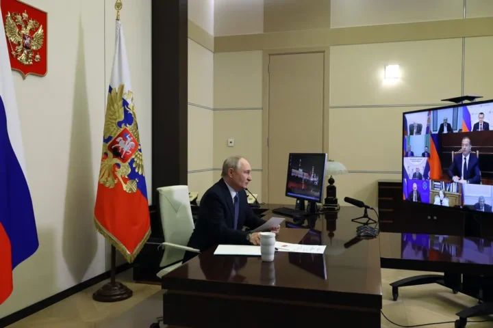 Vladimir Putin seated at a desk in front of Russian flags, taking part in a video meeting at Novo-Ogaryovo