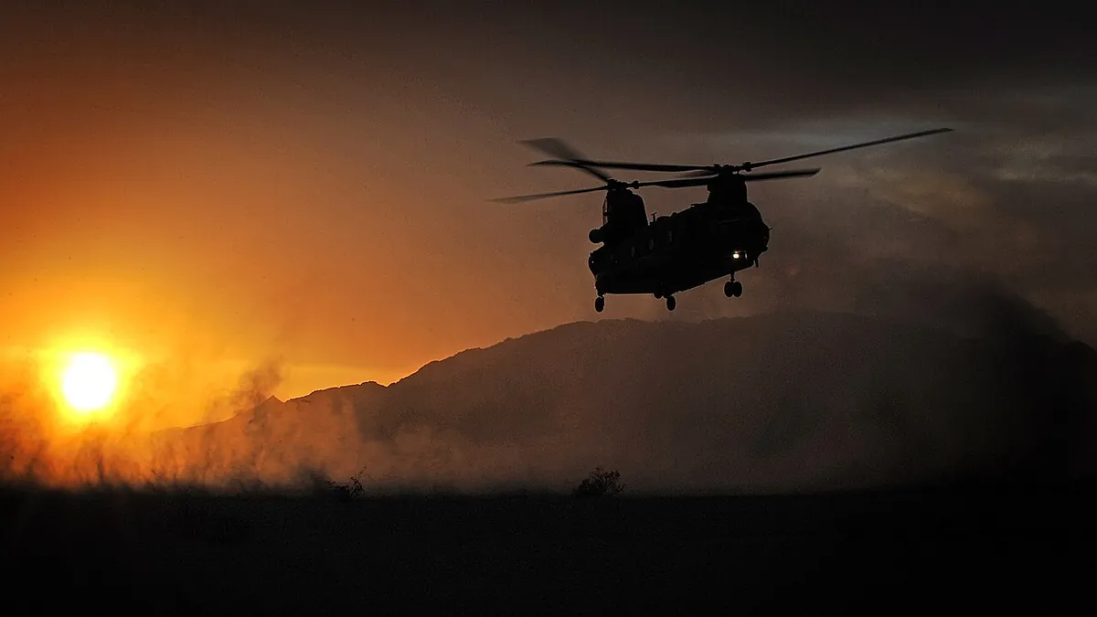 Silhouette of an RAF Chinook helicopter hovering at sunset over hills in a cloud of dust