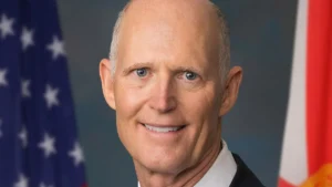 Senator Rick Scott in a dark suit and light blue tie in front of the American flag