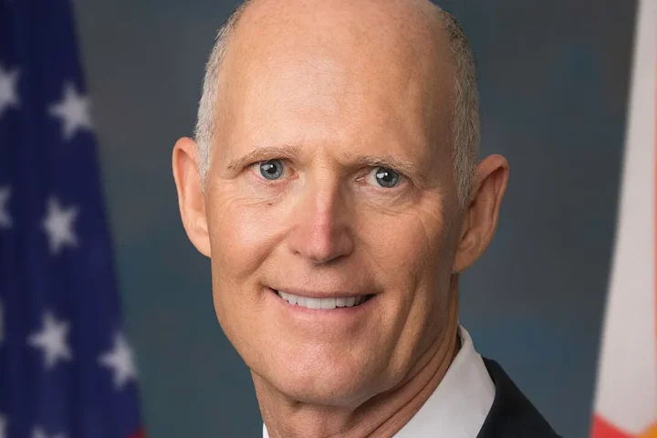 Senator Rick Scott in a dark suit and light blue tie in front of the American flag