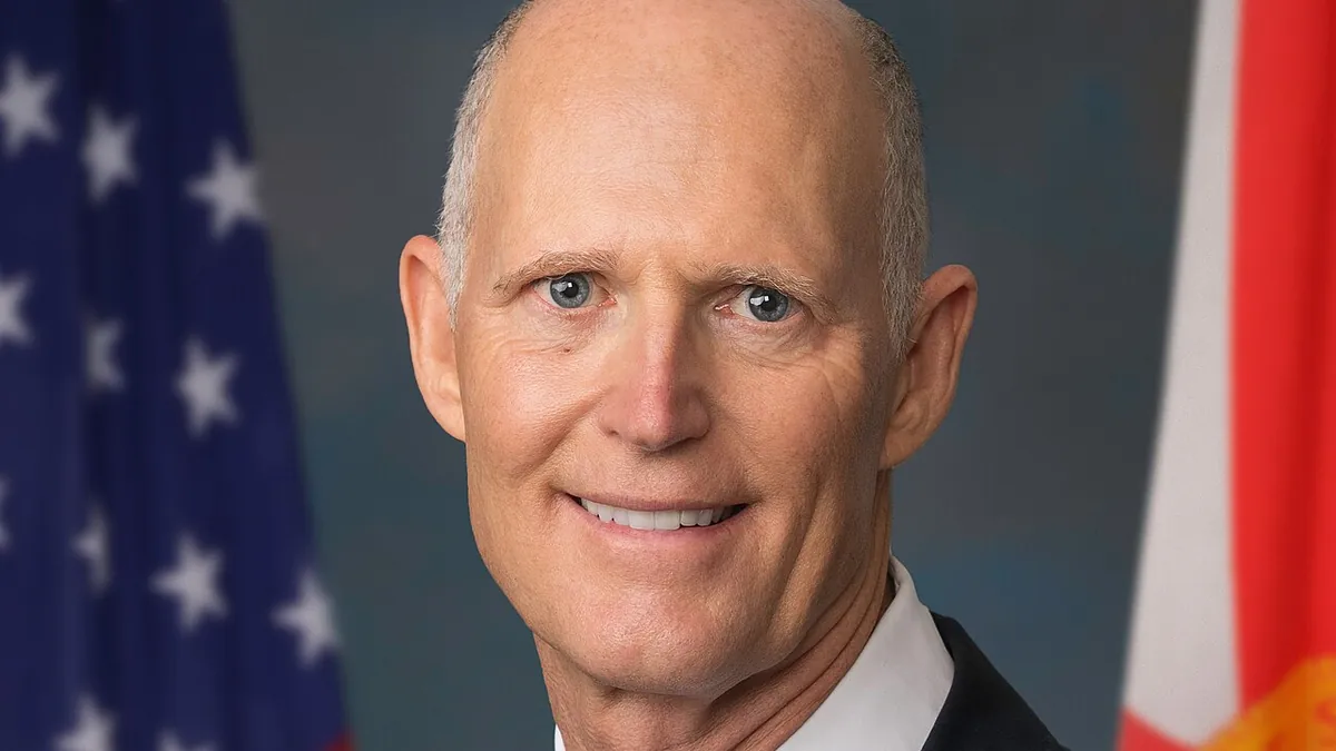 Senator Rick Scott in a dark suit and light blue tie in front of the American flag