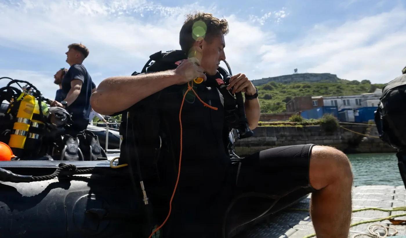 A Royal Navy diver in a wetsuit adjusts his breathing equipment beside an inflatable boat at Portland Port, with a green hillside behind