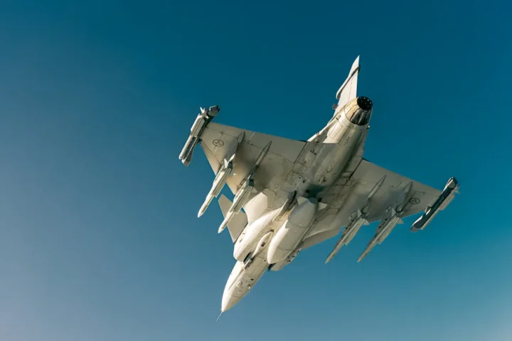 A Saab Gripen fighter seen from below in flight, carrying missiles and drop tanks