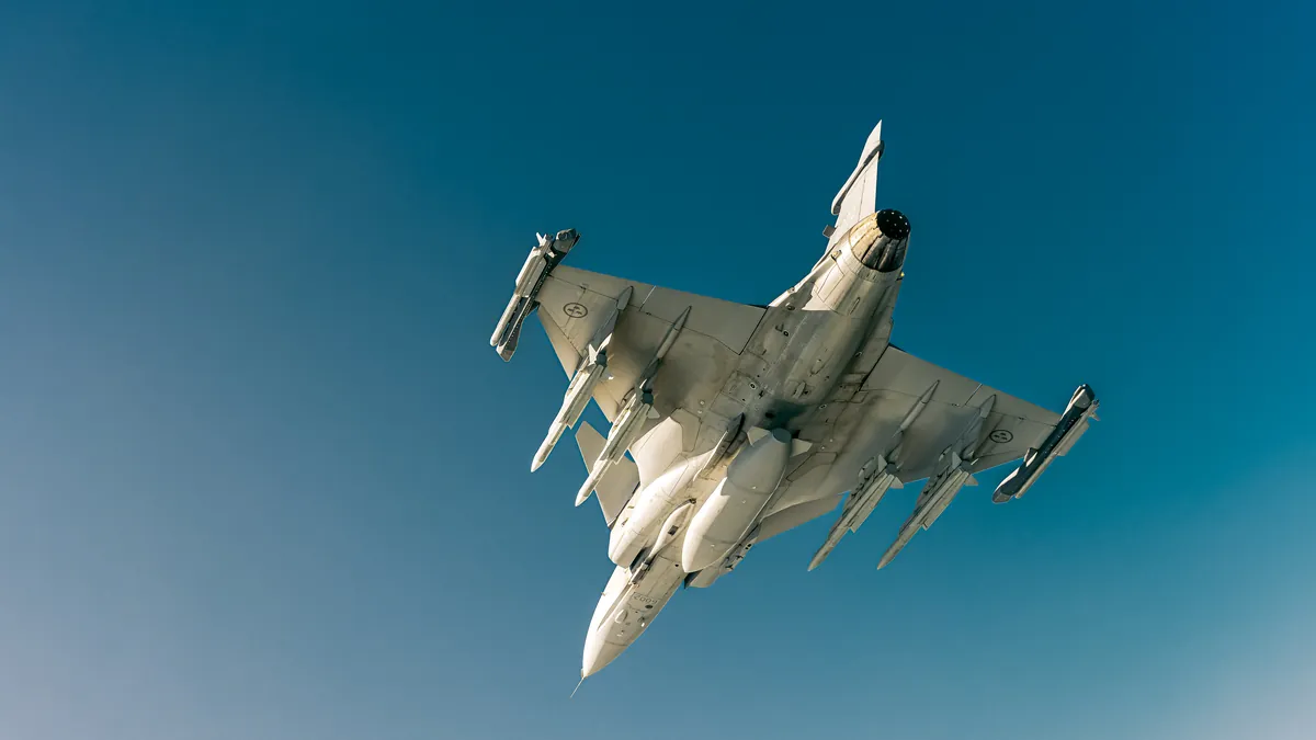 A Saab Gripen fighter seen from below in flight, carrying missiles and drop tanks