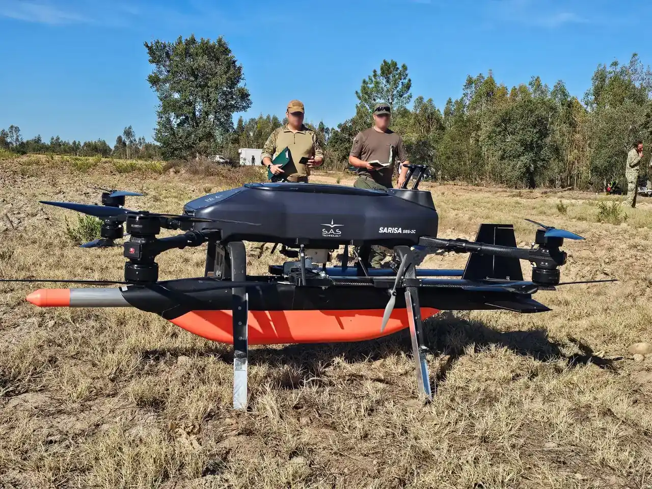The SARISA II multirotor UCAV carrying an AHM-2XR under its fuselage on grass, with two operators standing behind it