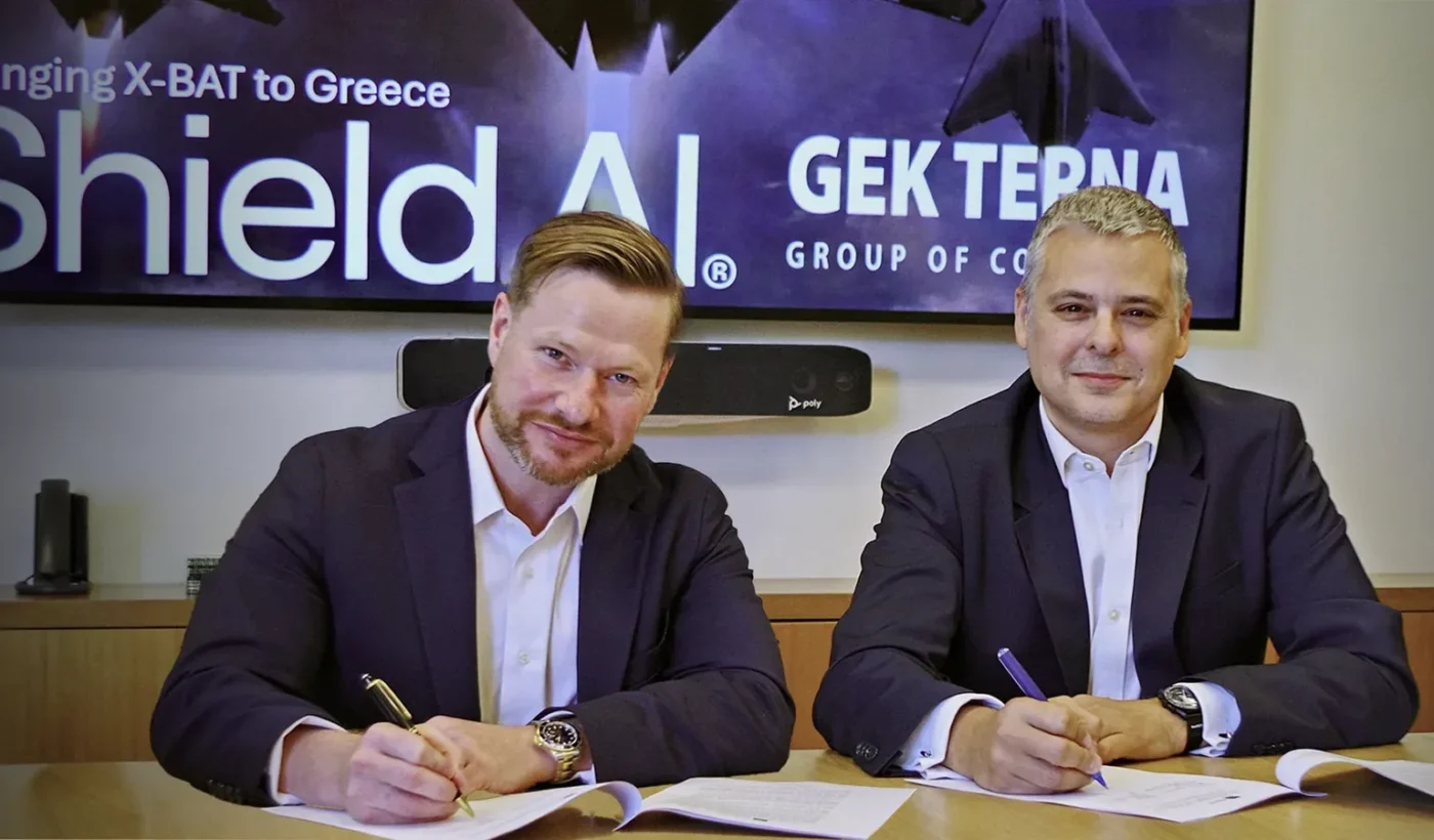 Two men in dark suits sign documents at a table in front of a screen reading Bringing X-BAT to Greece, with the Shield AI and GEK TERNA logos