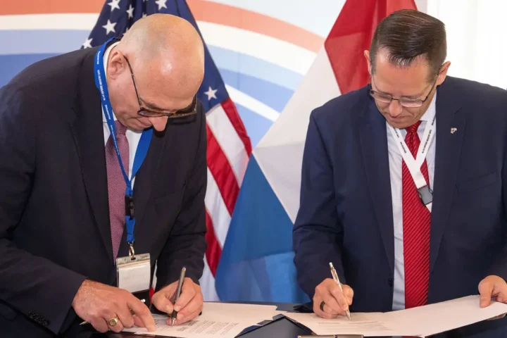 Two men sign documents in front of US and Dutch flags at the NL-US Defense Industry Days