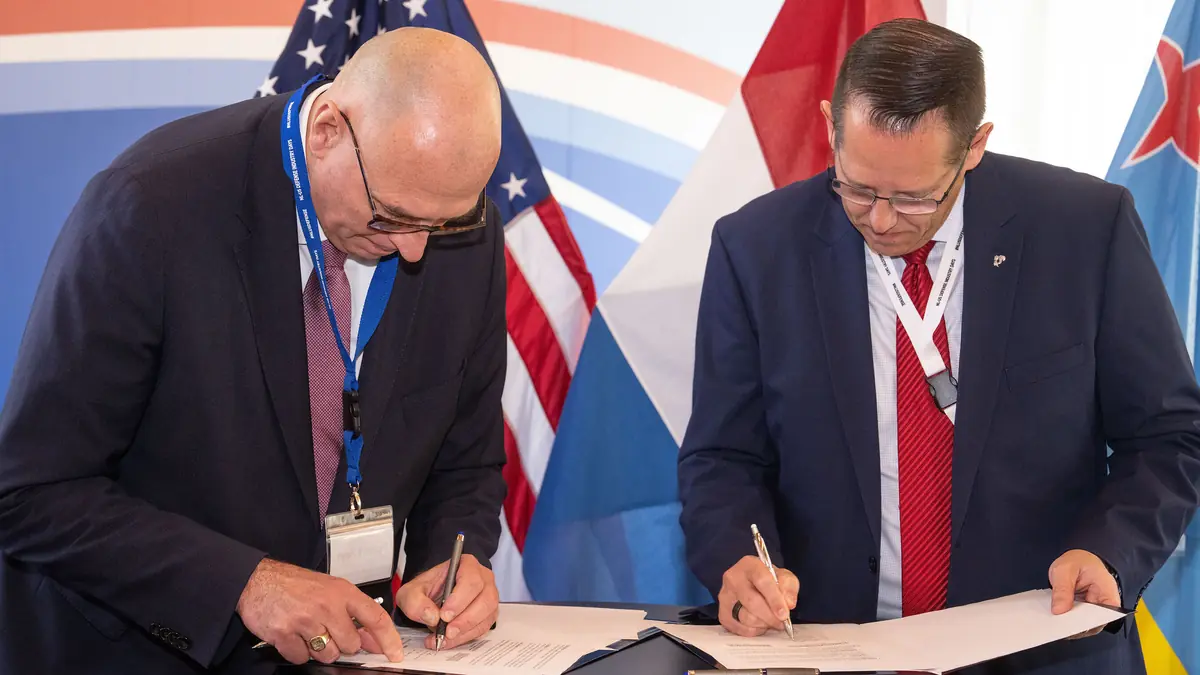 Two men sign documents in front of US and Dutch flags at the NL-US Defense Industry Days