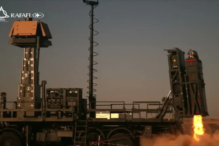The SPYDER AIO air-defence vehicle at dusk with its radar mast raised, firing a missile from the rear launcher with flame and dust at the base