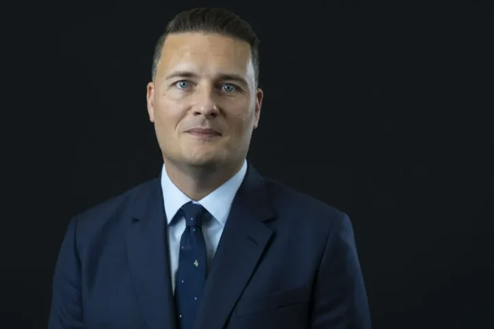 Portrait of UK Defence Secretary Wes Streeting in a dark suit and blue tie against a black background