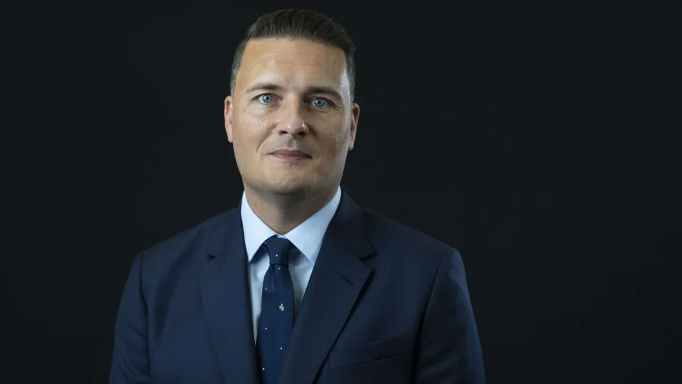 Portrait of UK Defence Secretary Wes Streeting in a dark suit and blue tie against a black background
