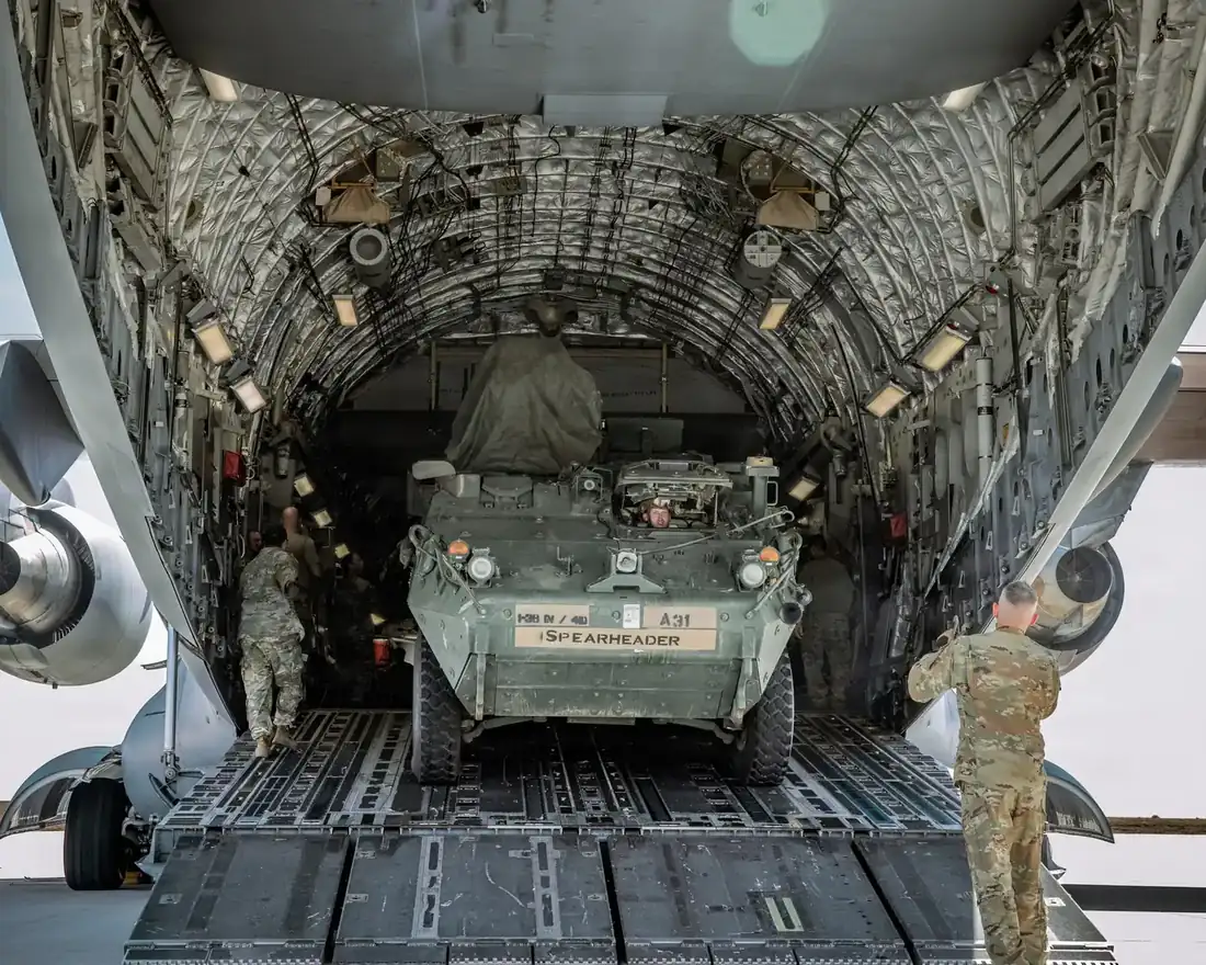 A Stryker with soldiers beside it inside the cargo hold of a US Air Force C-17 Globemaster III, seen from the rear ramp