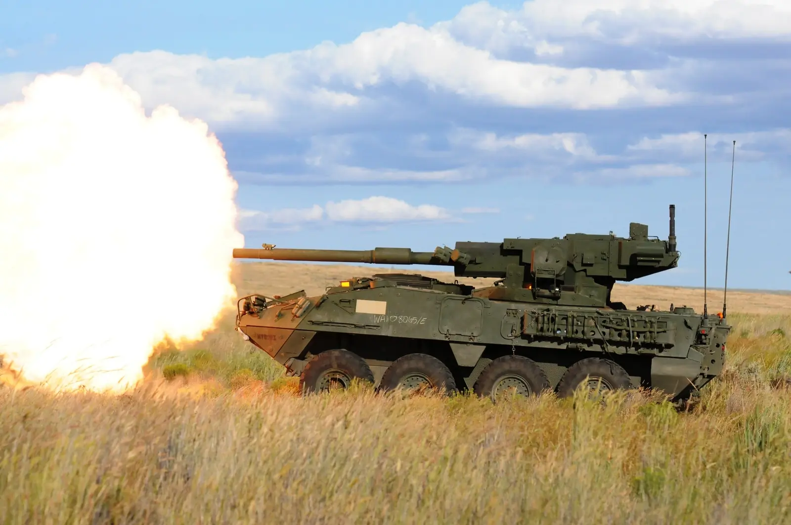 A Stryker M1128 Mobile Gun System firing its 105 mm gun on open grassland, with a large cloud of fire at the muzzle