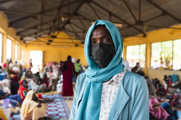 A woman in a face mask stands in a large hall where many people sit on mats