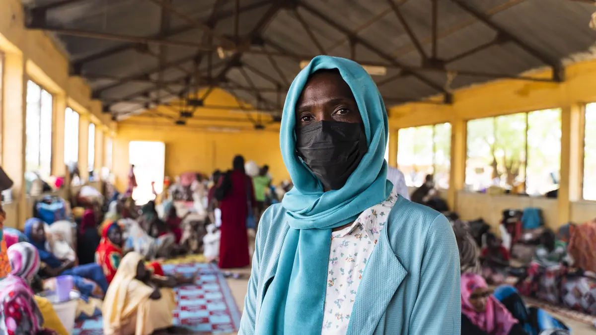 A woman in a face mask stands in a large hall where many people sit on mats