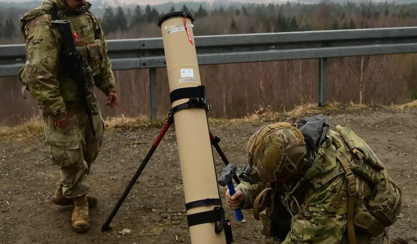 Two US Army soldiers setting up the tan launch tube of a Switchblade 600 loitering munition on a tripod at a training area