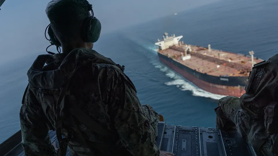 A U.S. service member looks out of a helicopter at a large tanker transiting the Strait of Hormuz