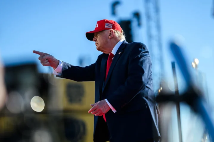 Donald Trump in a red cap and dark suit points with his right hand during a speech outdoors at Sparrows Point, Baltimore
