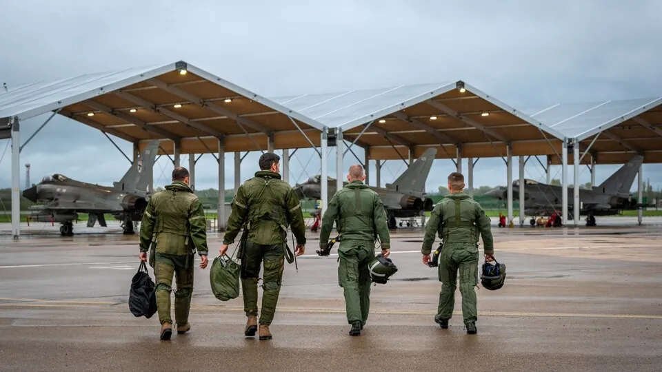 Four aircrew in flight suits walk across a wet apron towards RAF Typhoon fighters parked under open shelters