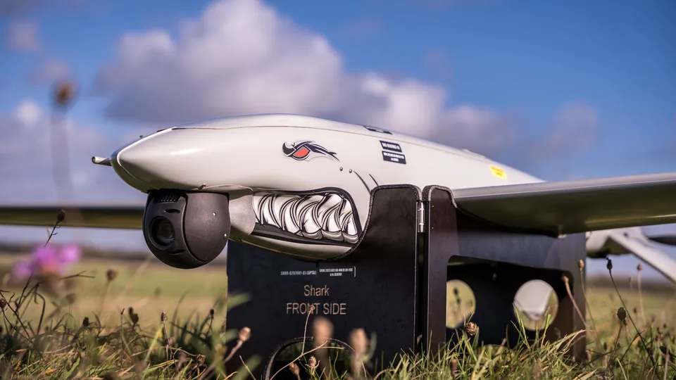 A grey uncrewed aircraft with a shark-mouth marking and a camera turret on a launch stand in grass under a blue sky