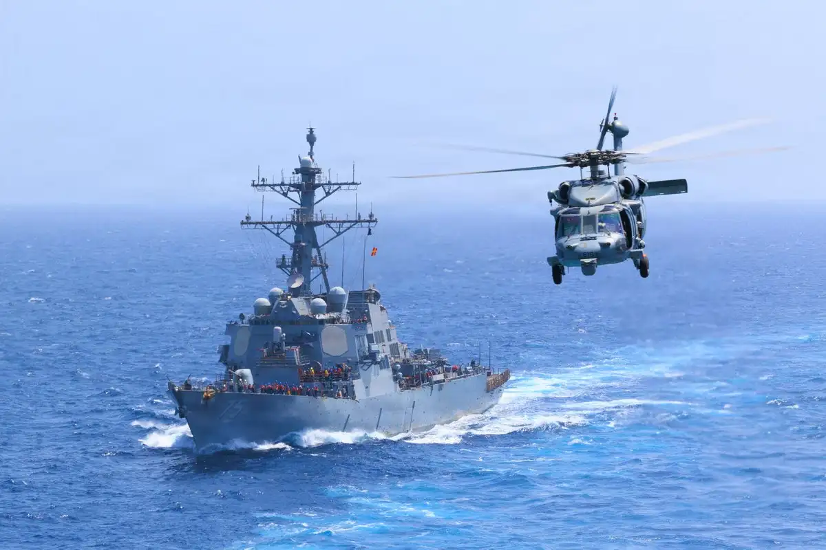 A US Navy guided-missile destroyer at sea with a Navy helicopter flying alongside it