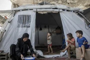 A woman prepares flour in a basin outside a tent in Gaza while three children sit and stand nearby