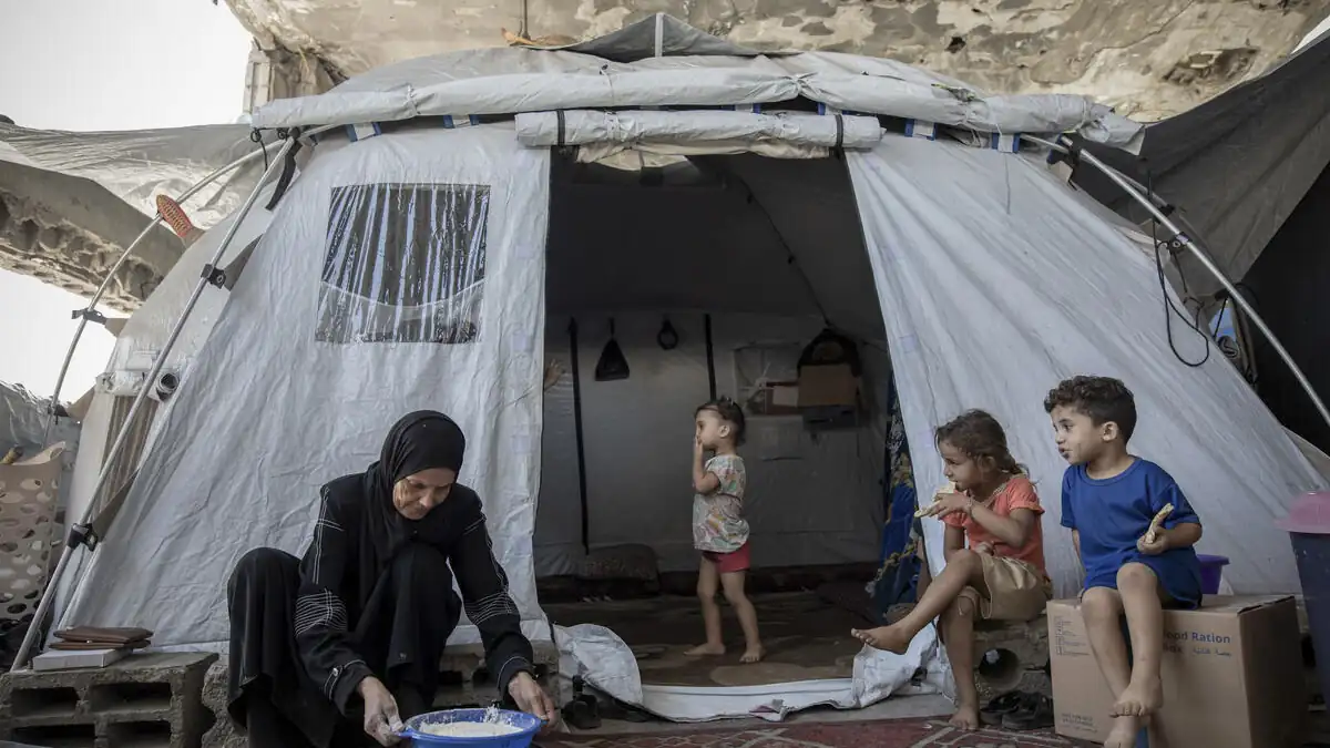 A woman prepares flour in a basin outside a tent in Gaza while three children sit and stand nearby